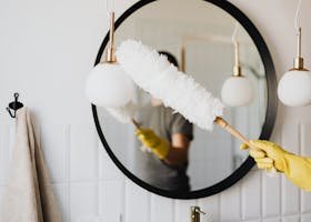 A person cleaning an elegant bathroom mirror with a fluffy duster and yellow gloves.